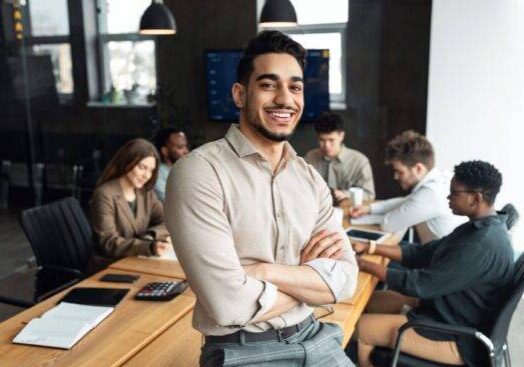 Successful Person. Portrait of confident smiling bearded businessman sitting leaning on desk in office, posing with folded arms and looking at camera, colleagues working in blurred background